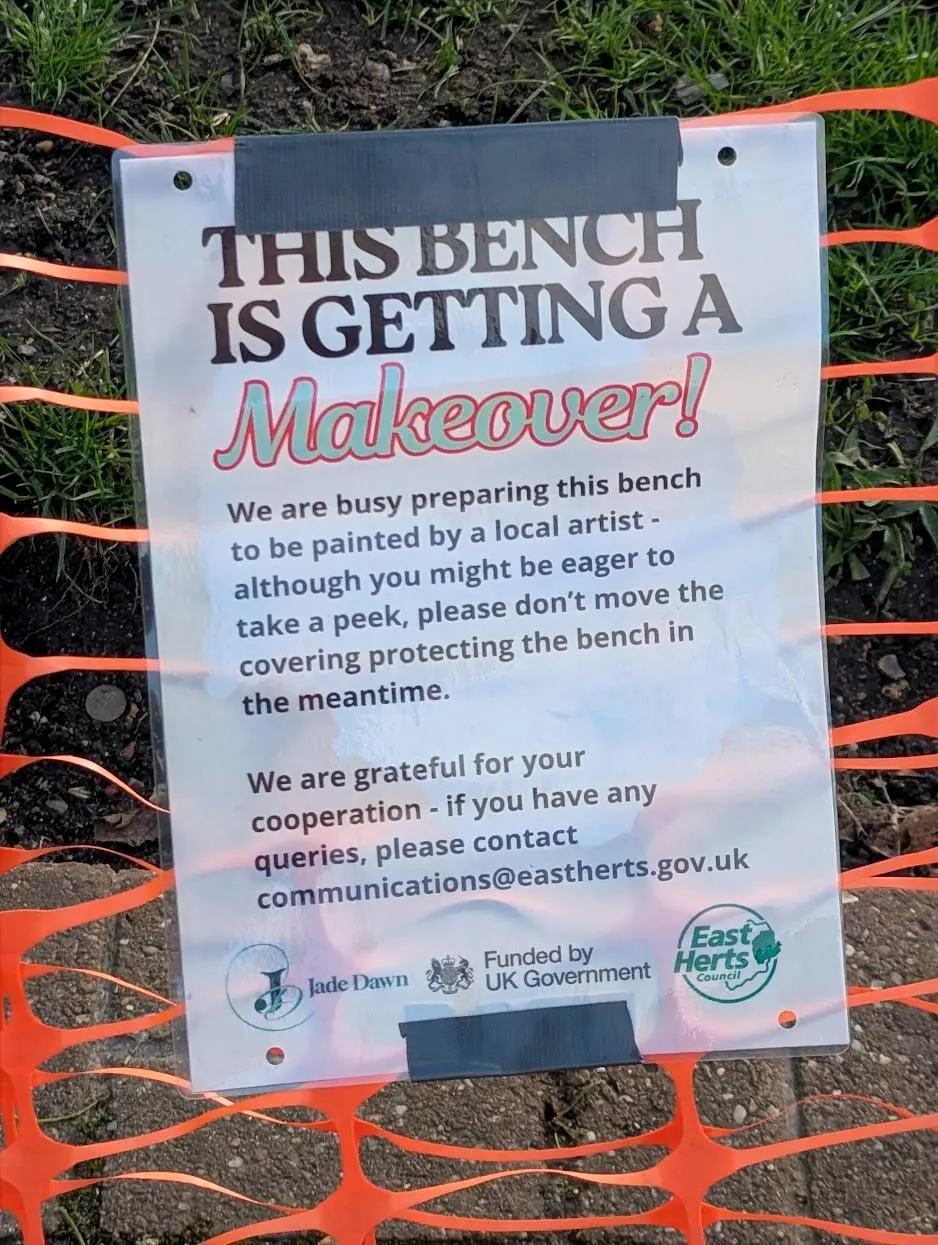 A poster explaining the bench is being repainted by a local artist. It says: This bench is getting a MAKEOVER! We are busy preparing this bench to be painted by a local artist - although you might be eager to take a peek, please dont remove the covering protecting the bench in the meantime. We are greatful for your cooperatiion - if you have any queries please contact communications@eastherts.gov.uk