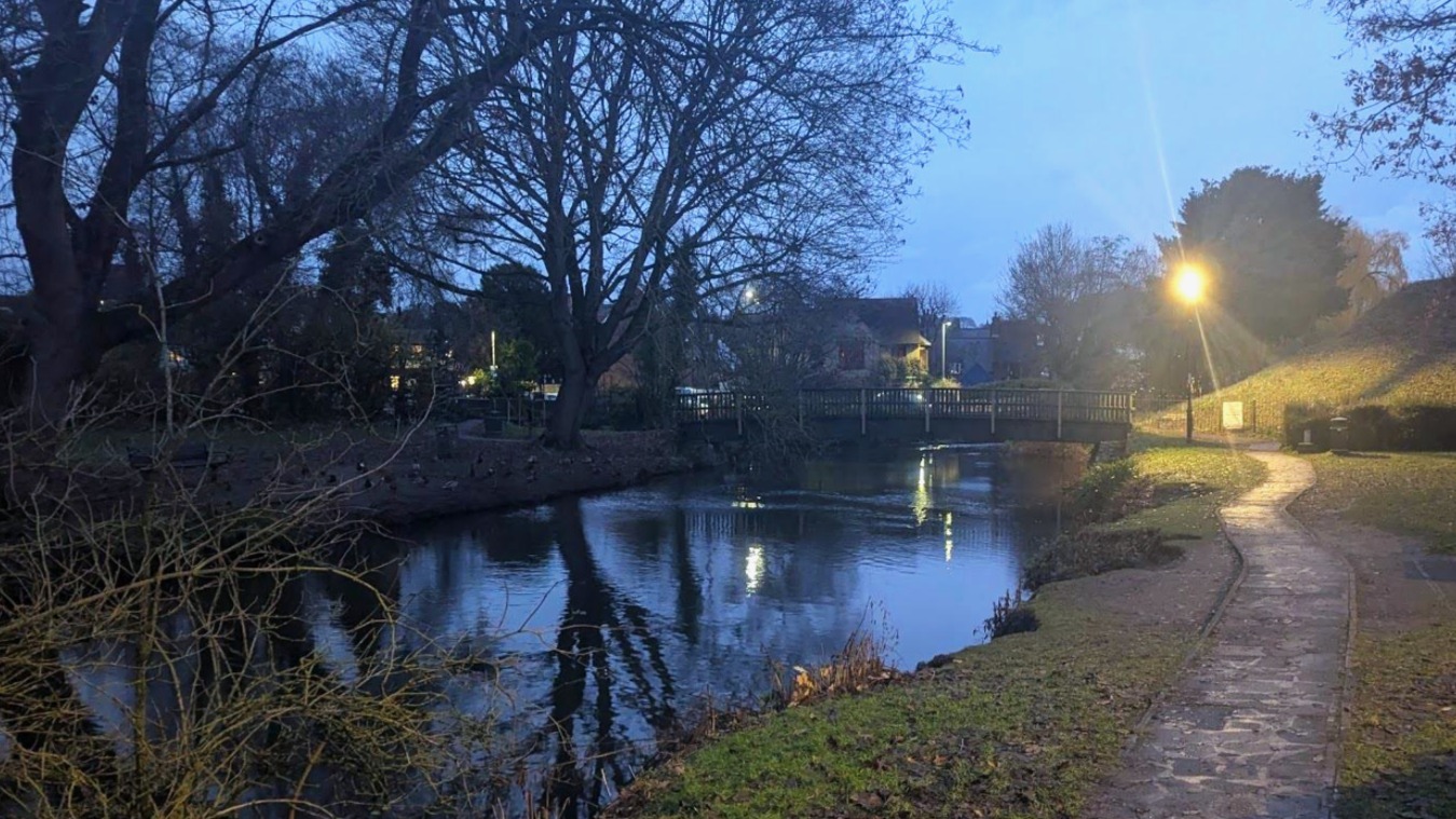 A landscape photo of a duck pond in early evening. The ducks are too cold for the water and sit on the side, a wooden bridge crosses the pond horizontally and a slippery path leads up to it.
